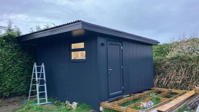A dark-clad garden building with a door and window, a deck frame laid in front